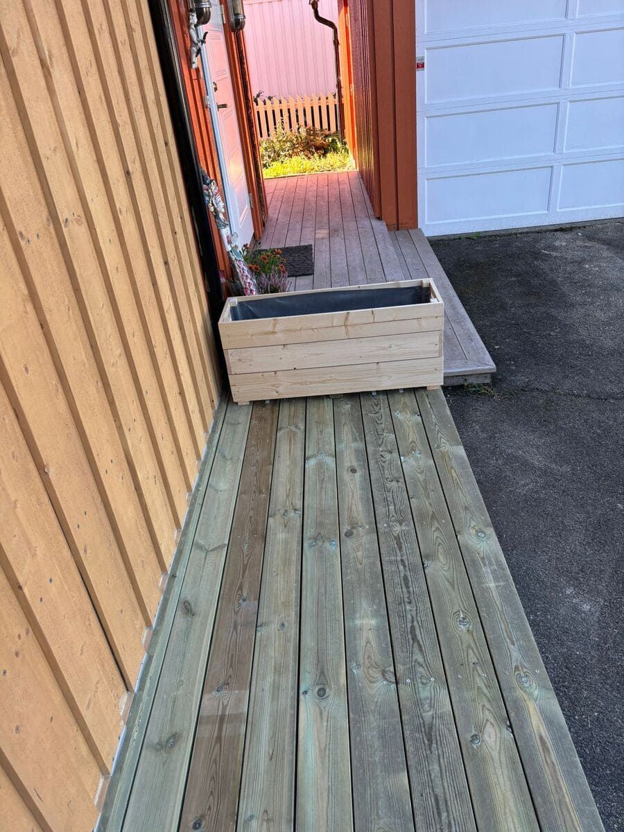 Rampa de madera que conduce a una puerta; jardinera de color claro en la rampa cerca de un edificio con puerta de garaje.