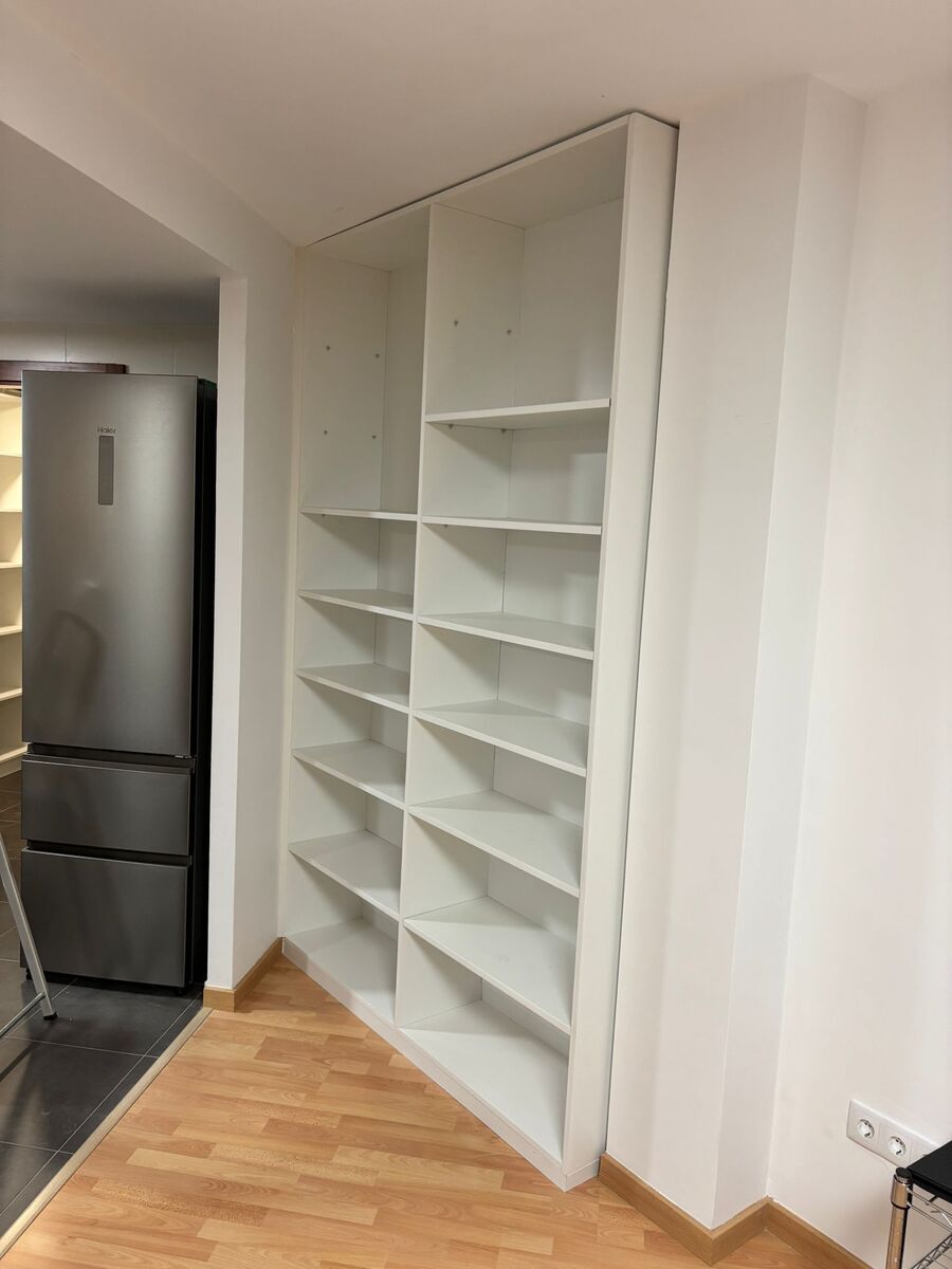 Estantería blanca empotrada en esquina, con varios estantes. Cerca se ve un refrigerador.