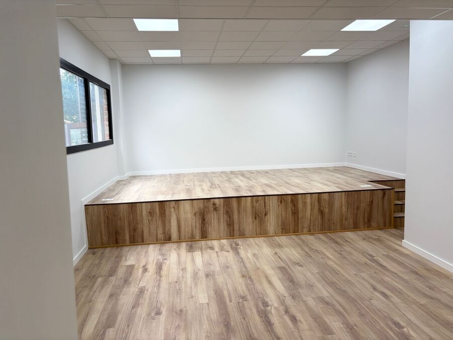 Habitación vacía con escenario y suelo de imitación madera, paredes blancas y una ventana.