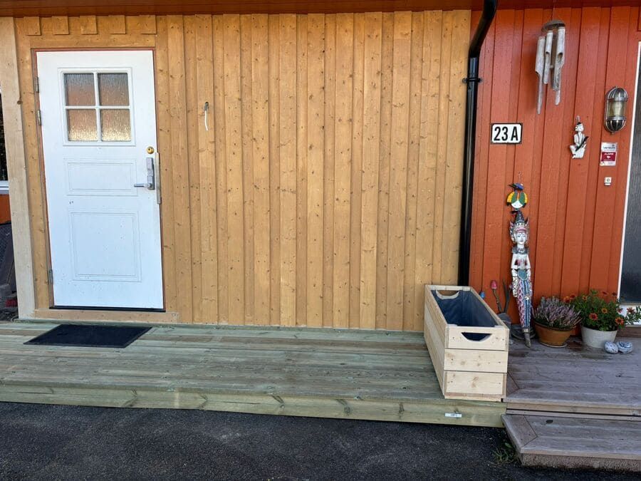 Exterior de edificio de madera con puerta blanca, terraza y pared naranja. Una jardinera de madera se encuentra en la terraza.