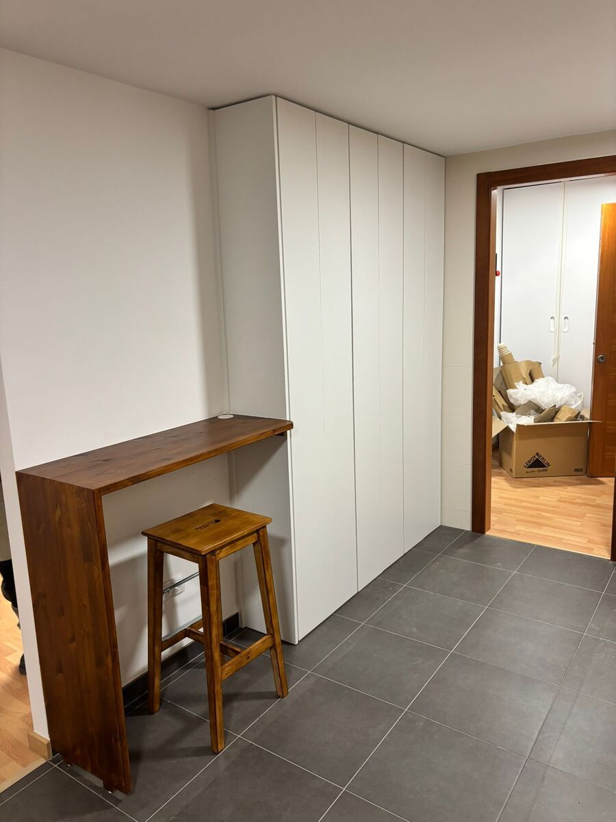 Un escritorio de madera con un taburete se encuentra junto a gabinetes de almacenamiento blancos en una habitación con piso de baldosas.