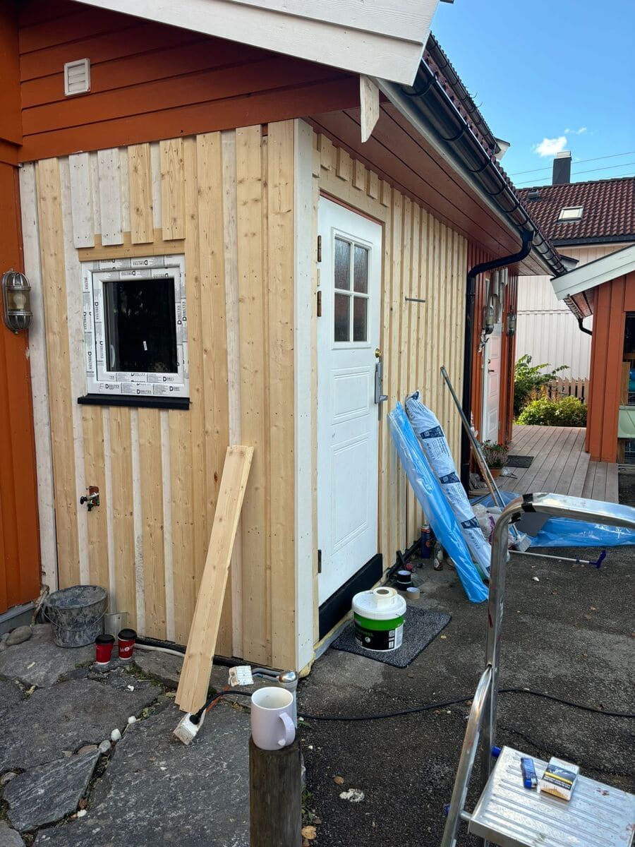 Exterior de un edificio pequeño con puerta y ventana blancas, que muestra revestimiento de madera sin terminar.