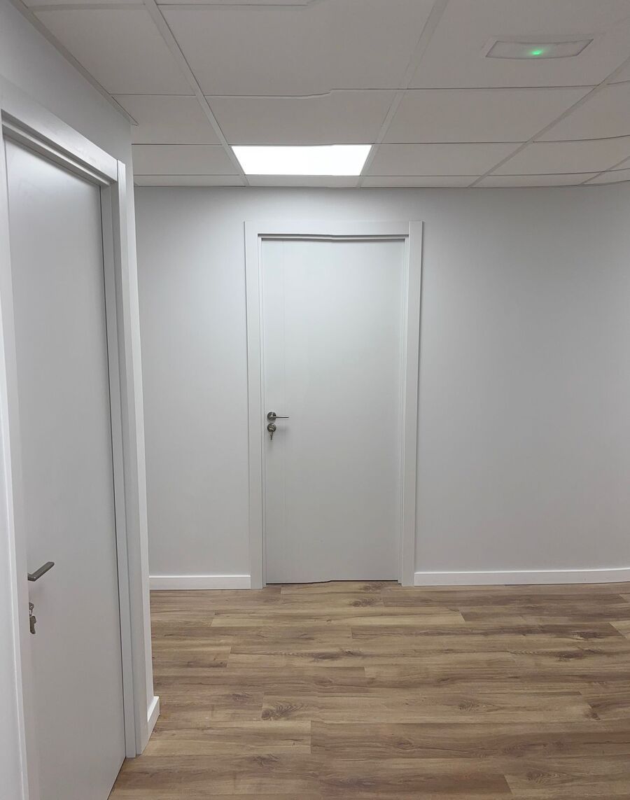 Un pasillo con paredes y puertas blancas, suelo de imitación madera clara y un panel de techo iluminado.
