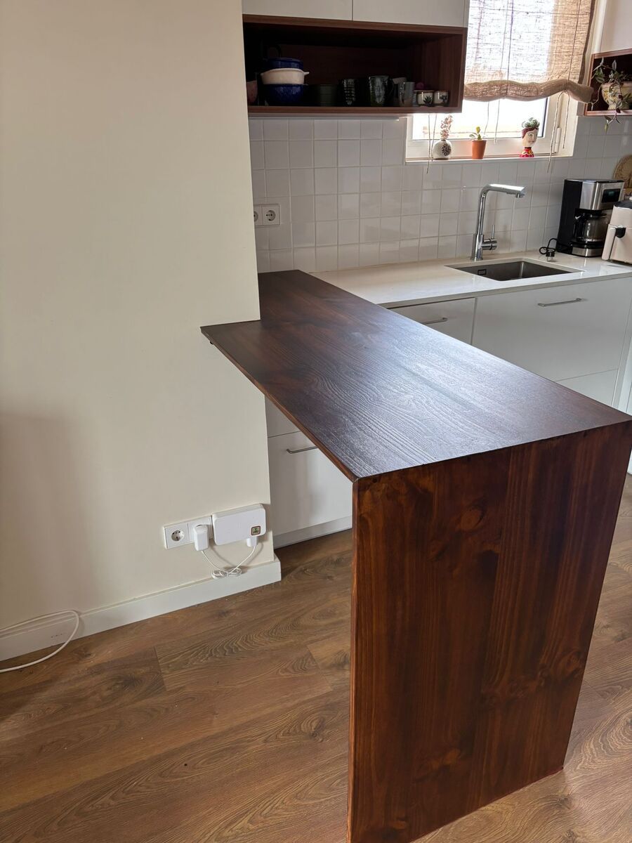 Encimera de cocina de madera oscura que sobresale de una pared blanca. Al fondo, se ven un fregadero y armarios.
