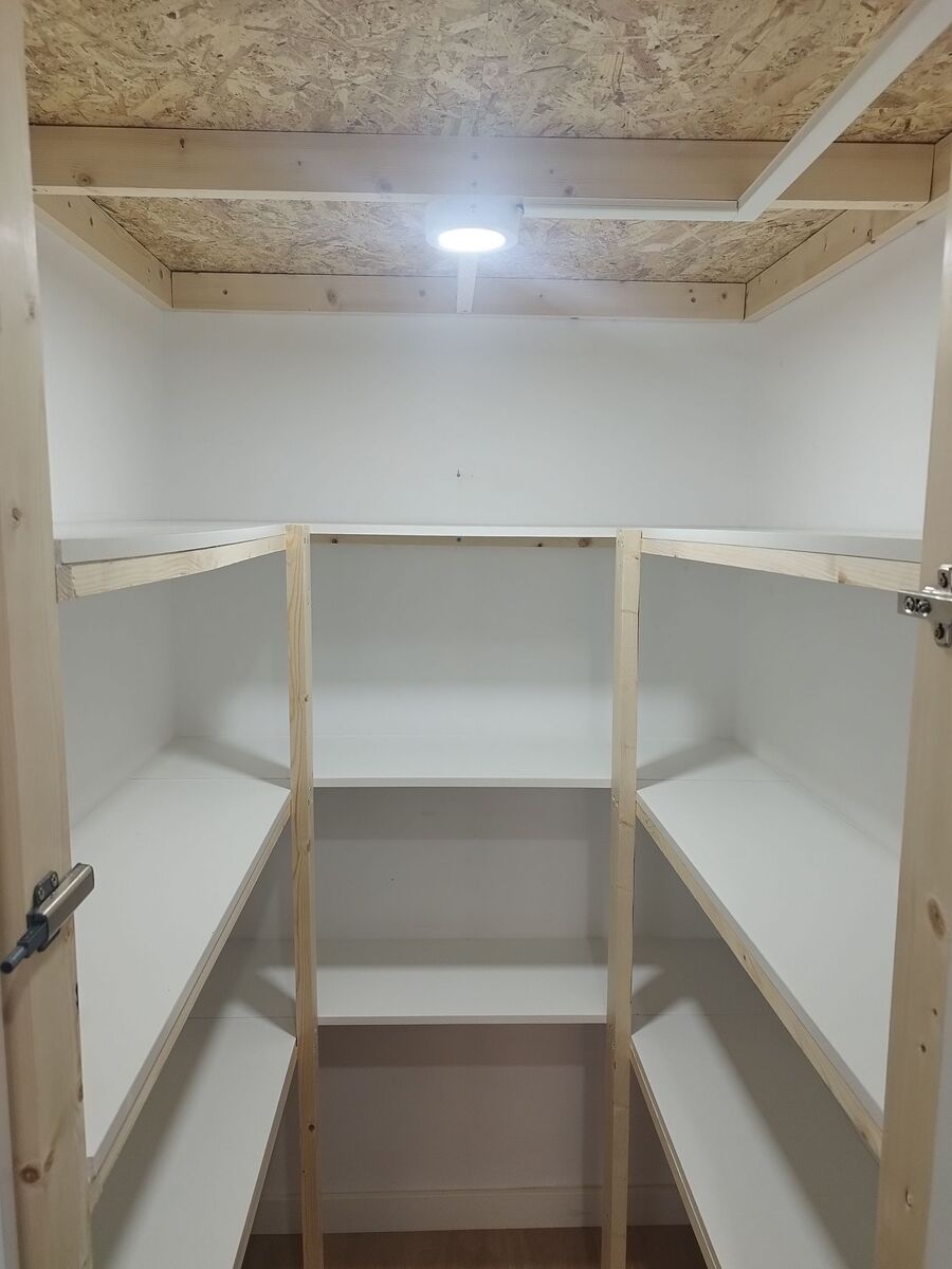 Despensa vacía con estantes blancos y una lámpara en el centro. Una moldura de madera clara rodea la abertura.