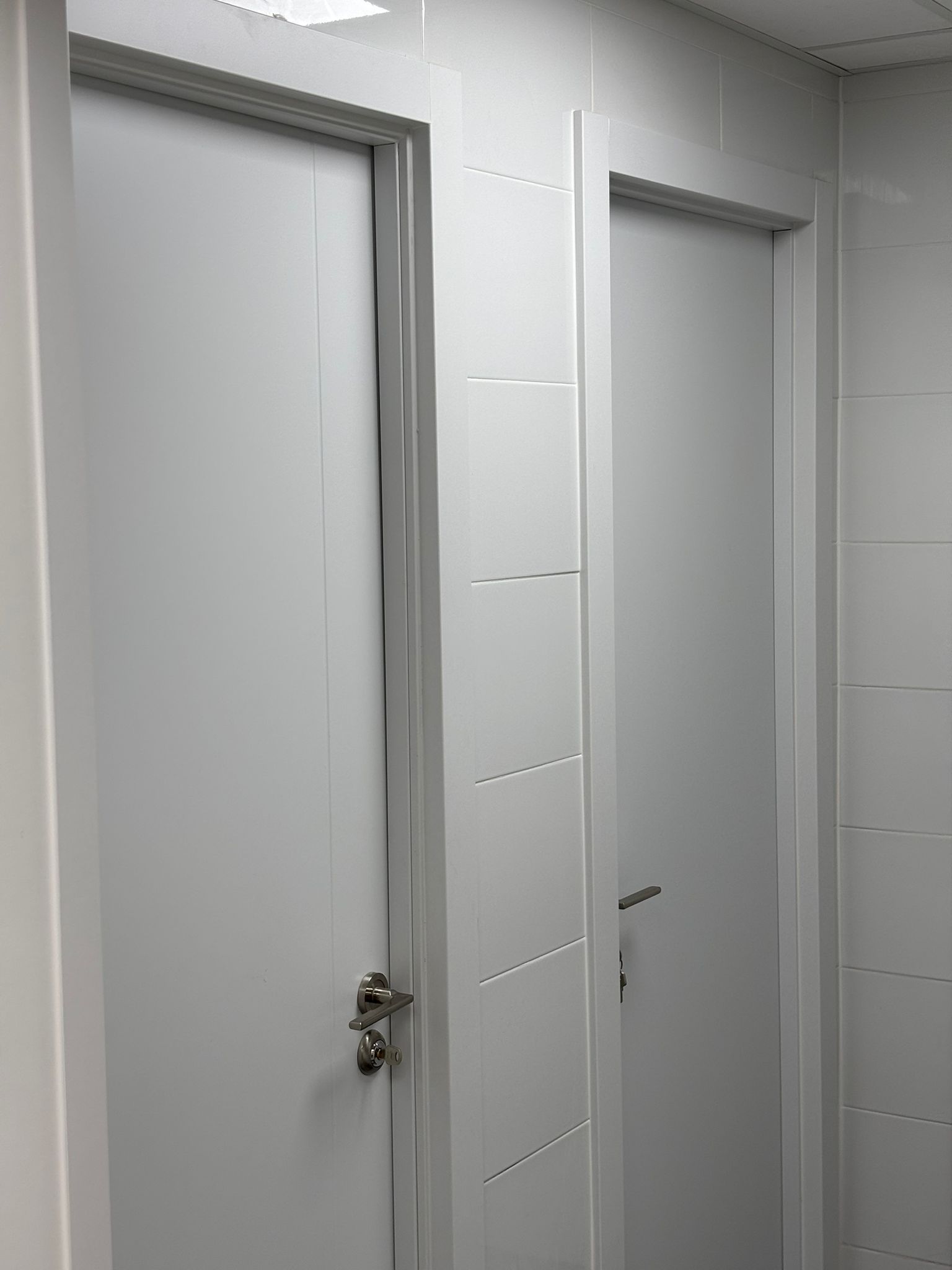 Dos puertas blancas en una habitación con azulejos blancos y marcos y manijas plateados.