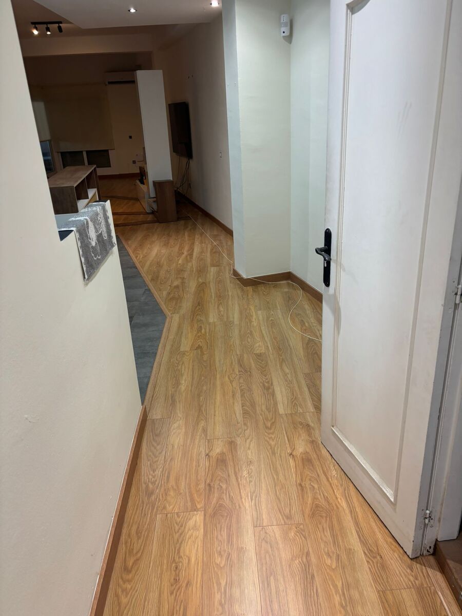 Pasillo con suelo imitación madera, paredes claras y puerta blanca con tirador negro a la derecha.