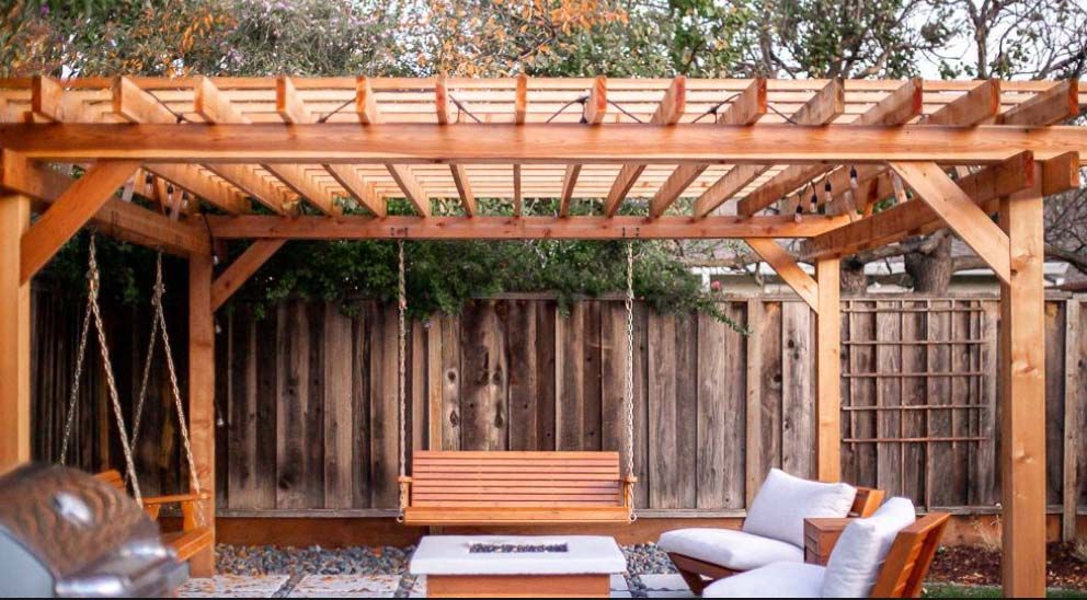 Pérgola de madera con columpio, banco y zona de estar en un patio trasero.