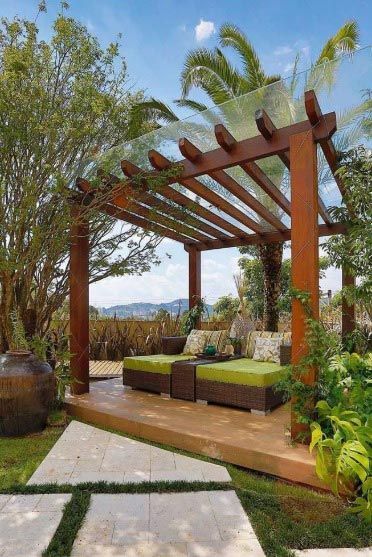 Pérgola de madera sobre zona de estar exterior con cojines verdes, rodeada de plantas y árboles.