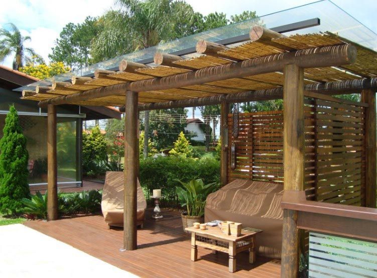 Pérgola de madera con terraza, zona de estar y techo de cristal. Está rodeada de plantas y un jardín.