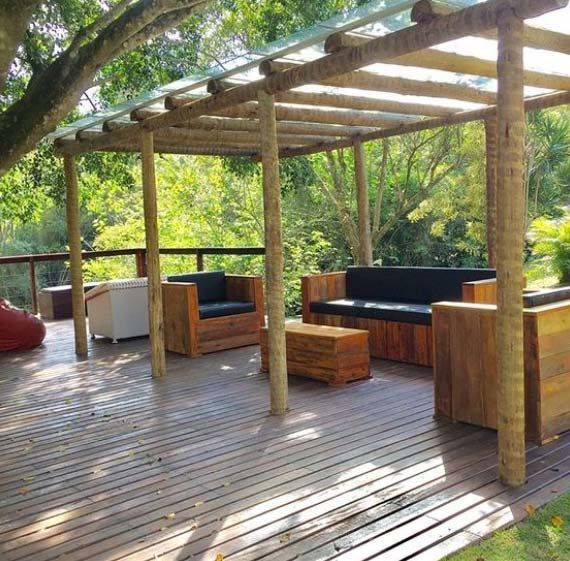 Terraza de madera con zona de estar bajo una pérgola. Incluye muebles de madera y un fondo verde frondoso.
