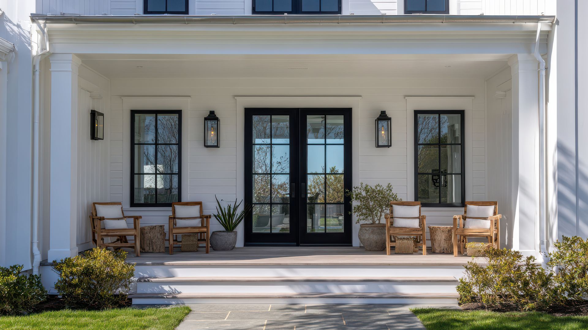 Le porche d'une maison blanche est orné d'une double porte noire, de deux ensembles de chaises en bois et d'un éclairage extérieur symétrique.