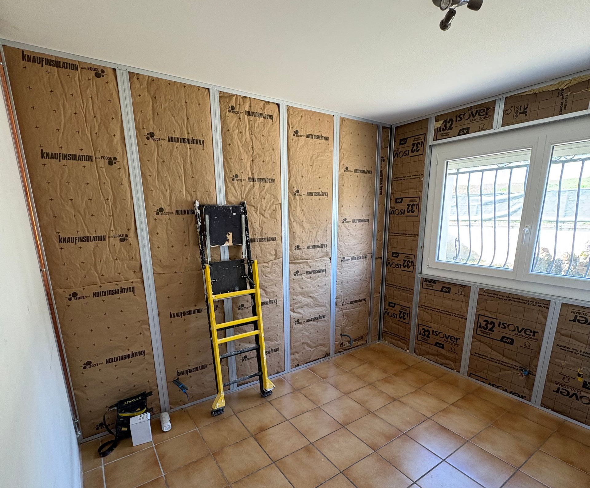 Une pièce intérieure en construction présente des montants muraux métalliques remplis de panneaux isolants bruns et un escabeau jaune.