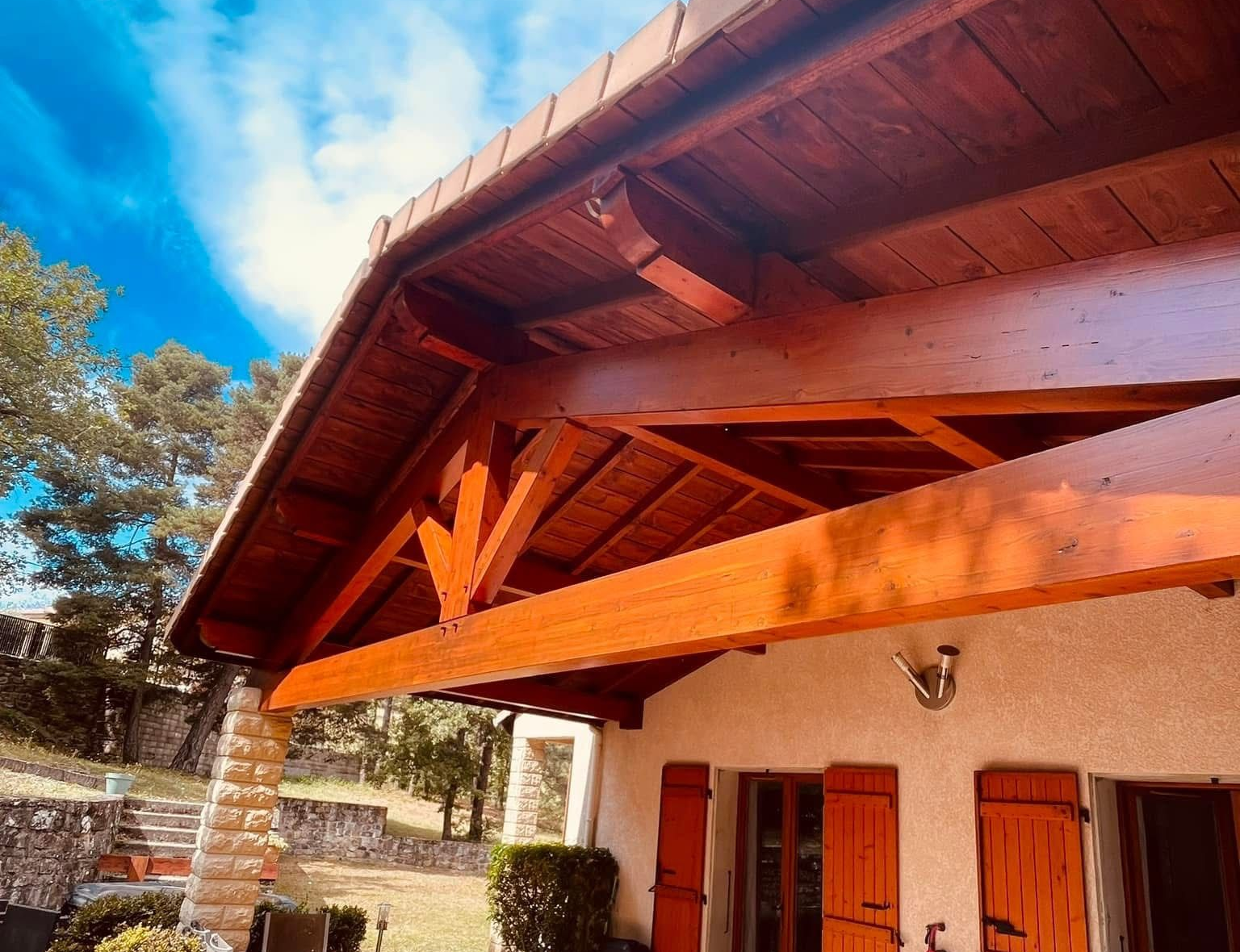 Terrasse d’une maison avec charpente en bois verni, volets en bois, mobilier extérieur et végétation environnante sous un ciel bleu partiellement nuageux.