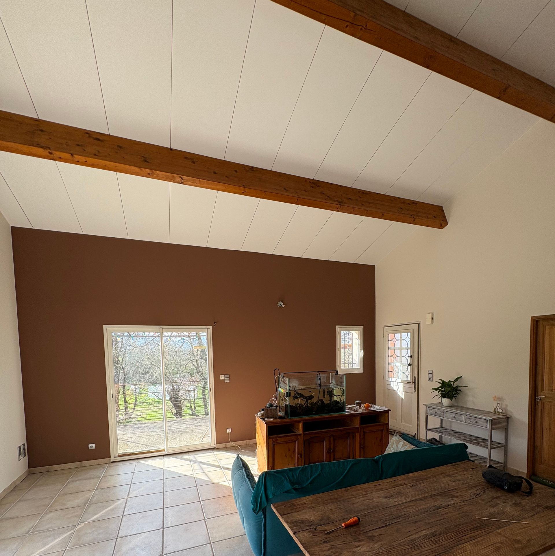 Un salon avec un plafond voûté, des poutres apparentes, un mur d'accent marron, un canapé turquoise et des portes coulissantes en verre.