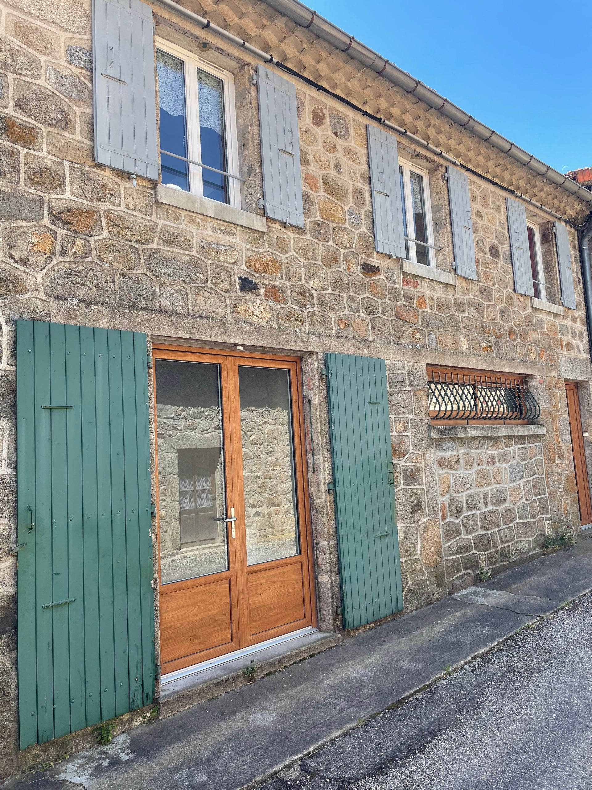 L'extérieur rustique d'une maison en pierre, avec une porte d'entrée aux volets verts et des fenêtres à l'étage aux volets gris, se détache sur un ciel bleu.