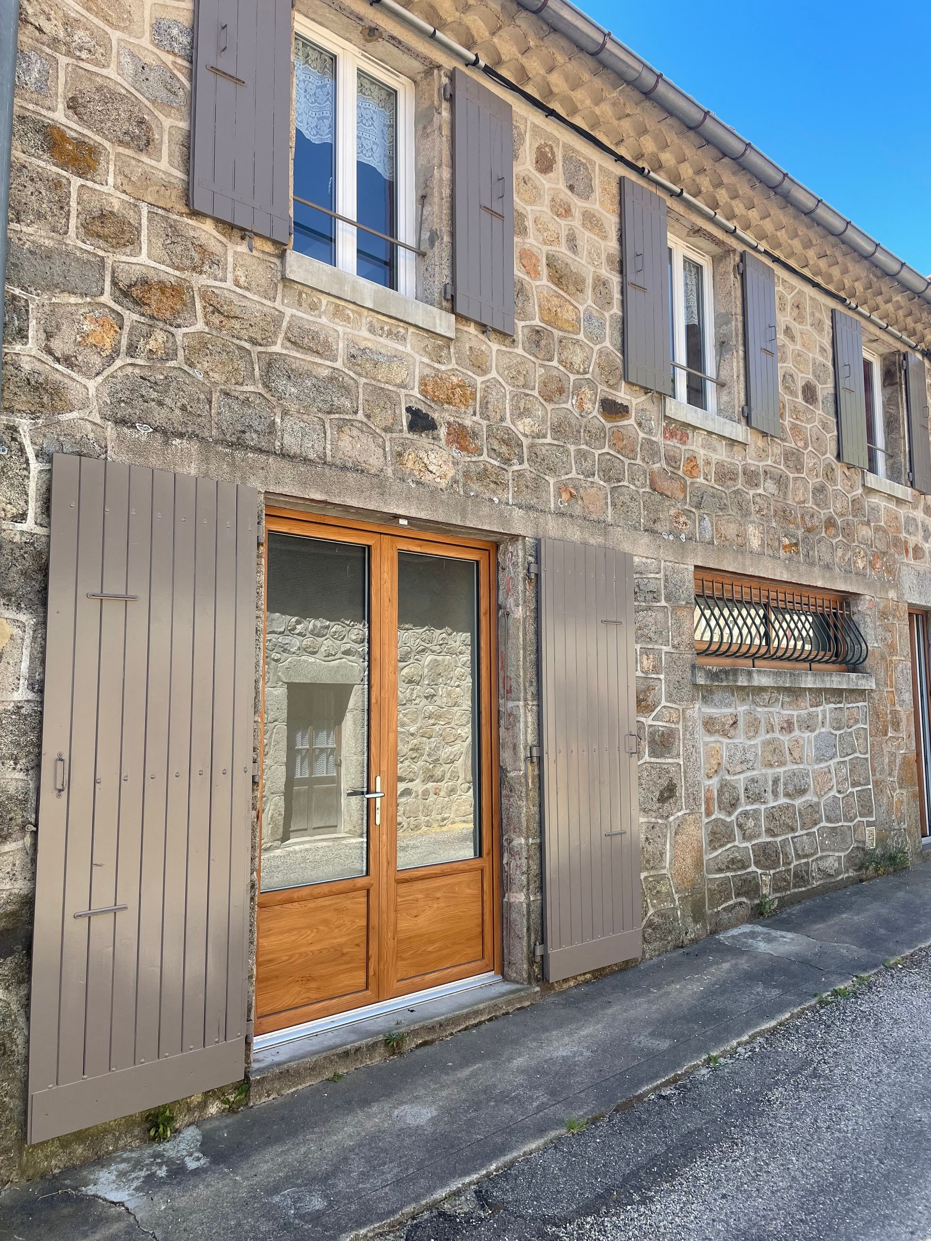 Maison en pierre de deux étages avec une porte d'entrée en bois, des volets en bois clair et plusieurs fenêtres.