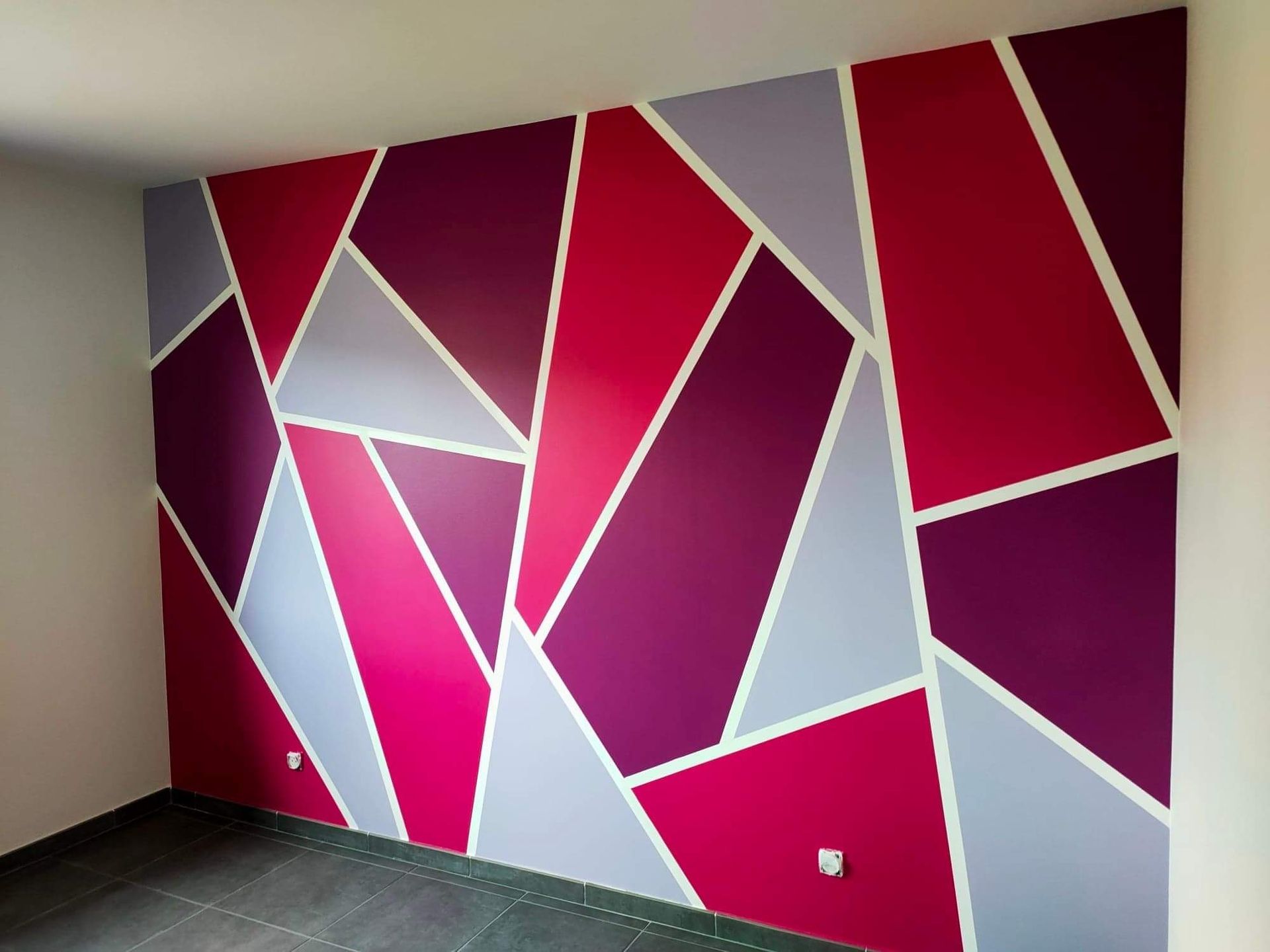 Une fresque murale géométrique présentant des formes angulaires et nettes peintes dans des tons de magenta, de violet et de gris clair.