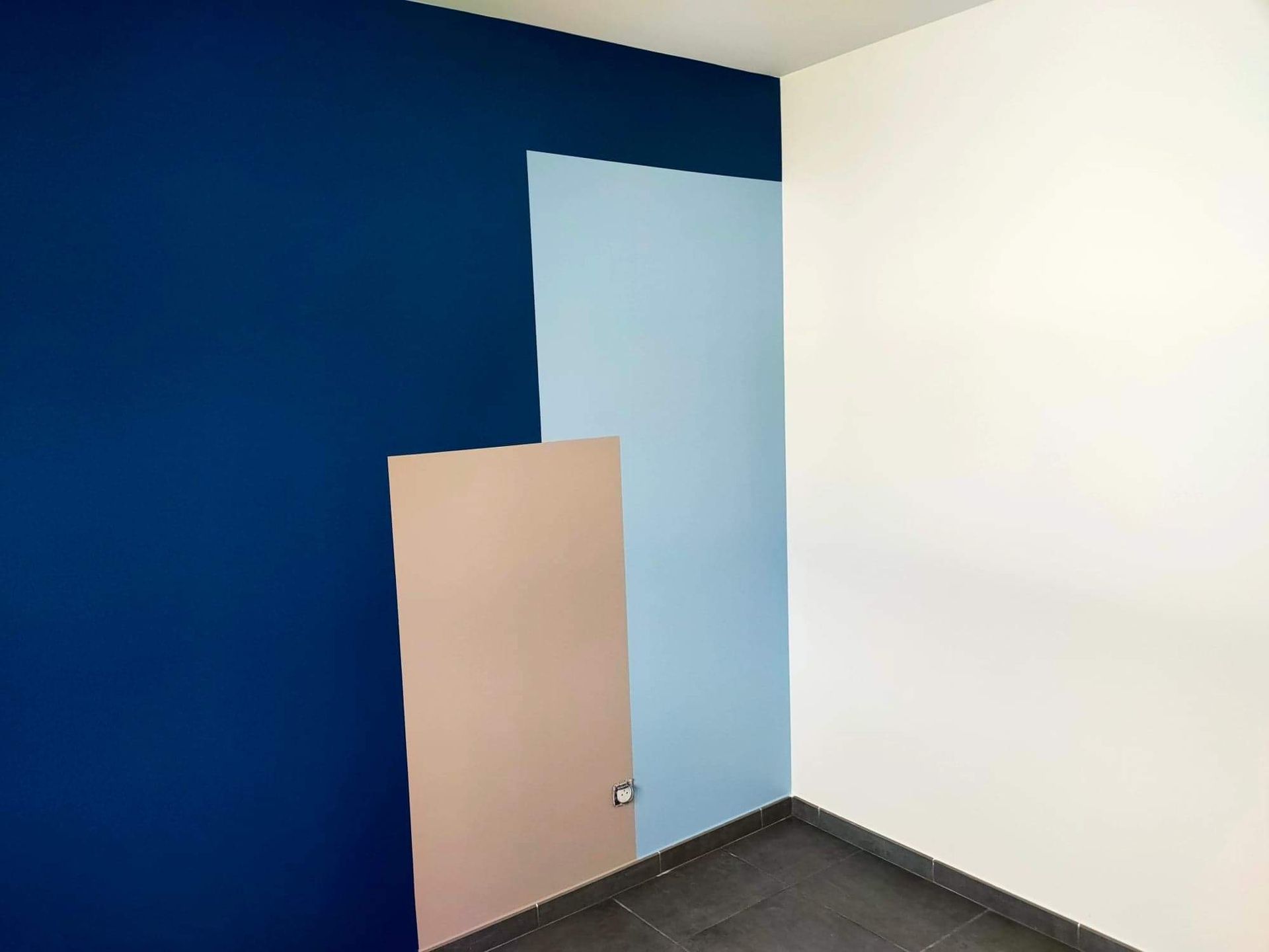 Mur intérieur peint avec trois aplats de couleurs contrastées : un bleu foncé, un bleu clair et un beige, créant une composition géométrique sur un sol en carrelage gris.
