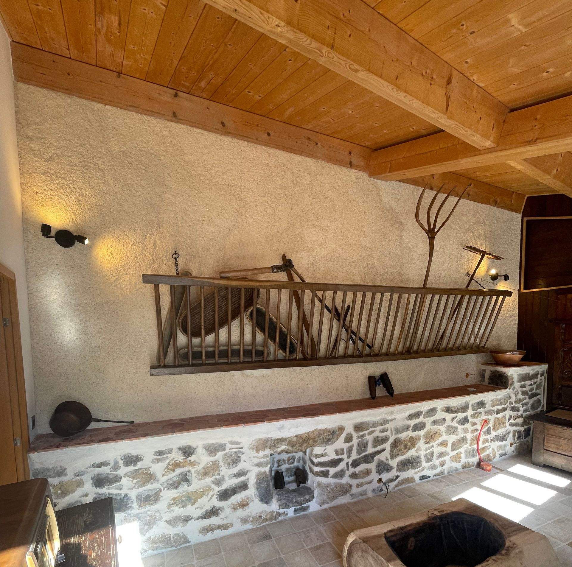 Un espace intérieur rustique présente un mur texturé avec un râtelier à foin et des outils agricoles anciens au-dessus d'une corniche en pierre et d'un plafond en bois.