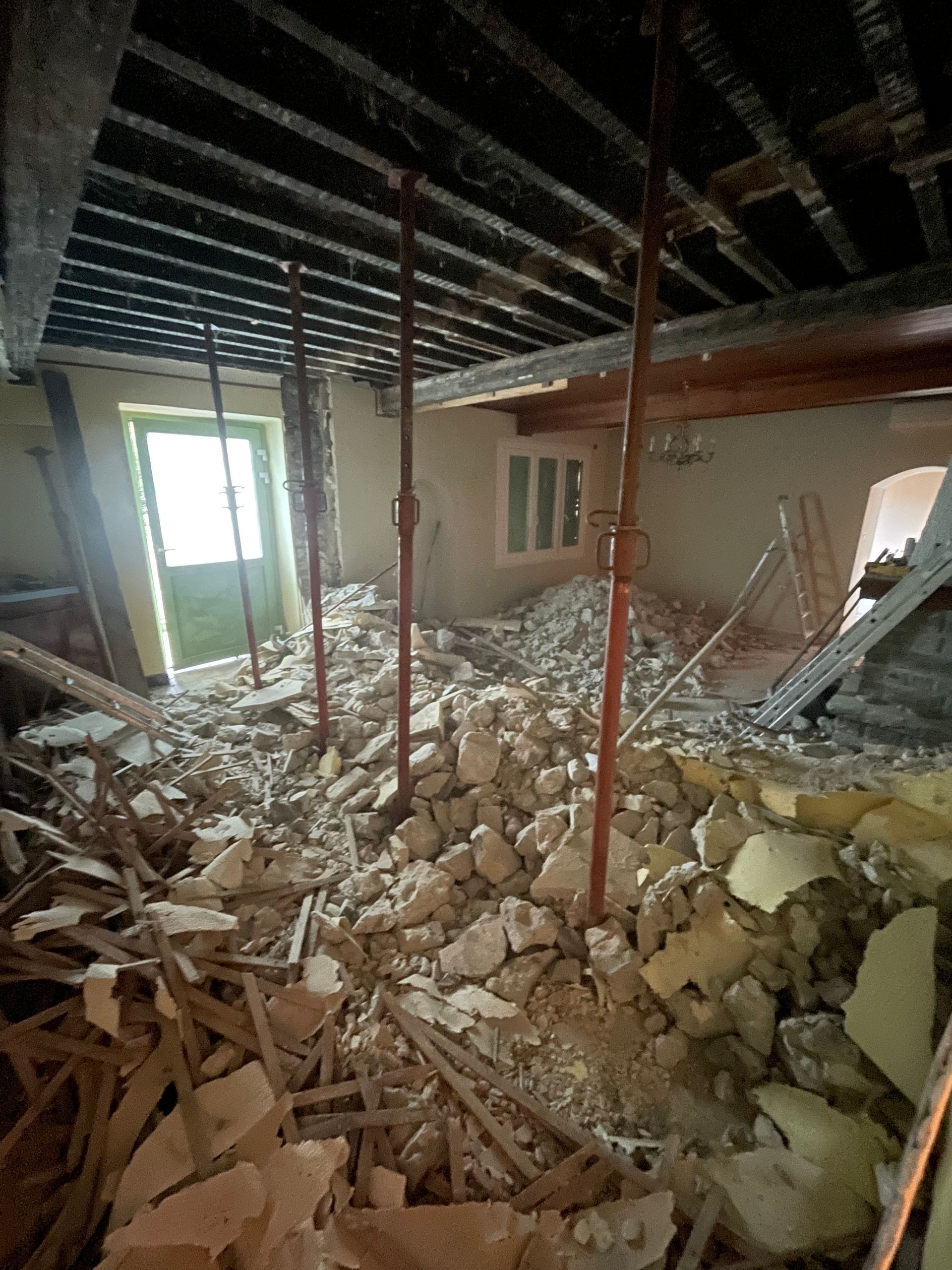 Un chantier de construction intérieur montrant des poutres de plafond apparentes, des vérins de soutien en métal rouge et un sol recouvert de débris.