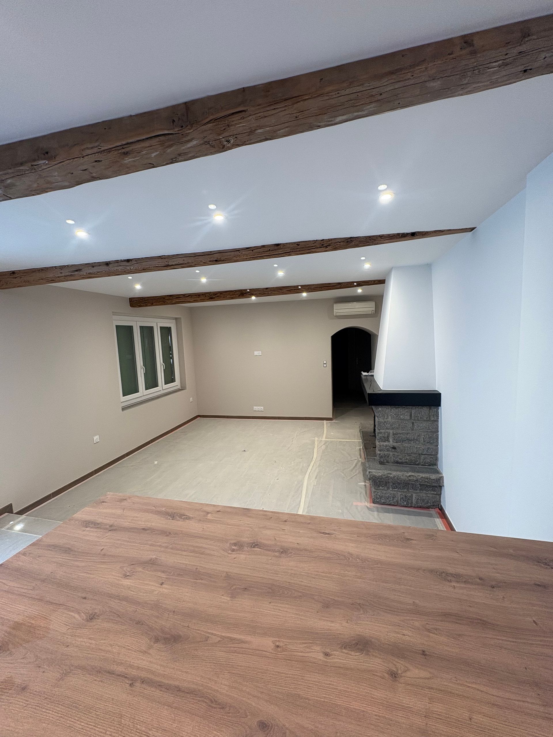 Une pièce spacieuse et moderne avec un sol clair, des poutres apparentes au plafond, un éclairage encastré et une cheminée en pierre.