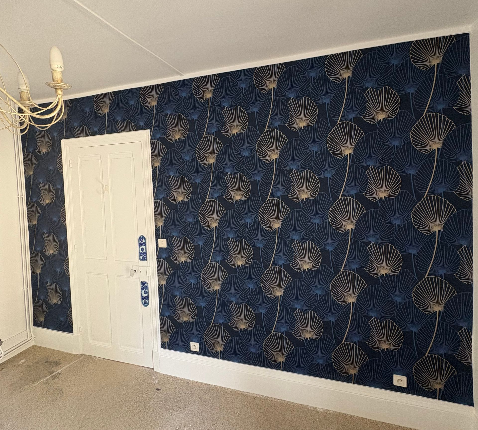 Une porte blanche encastrée dans un mur recouvert d'un papier peint bleu foncé orné d'un motif botanique doré en forme d'éventail.