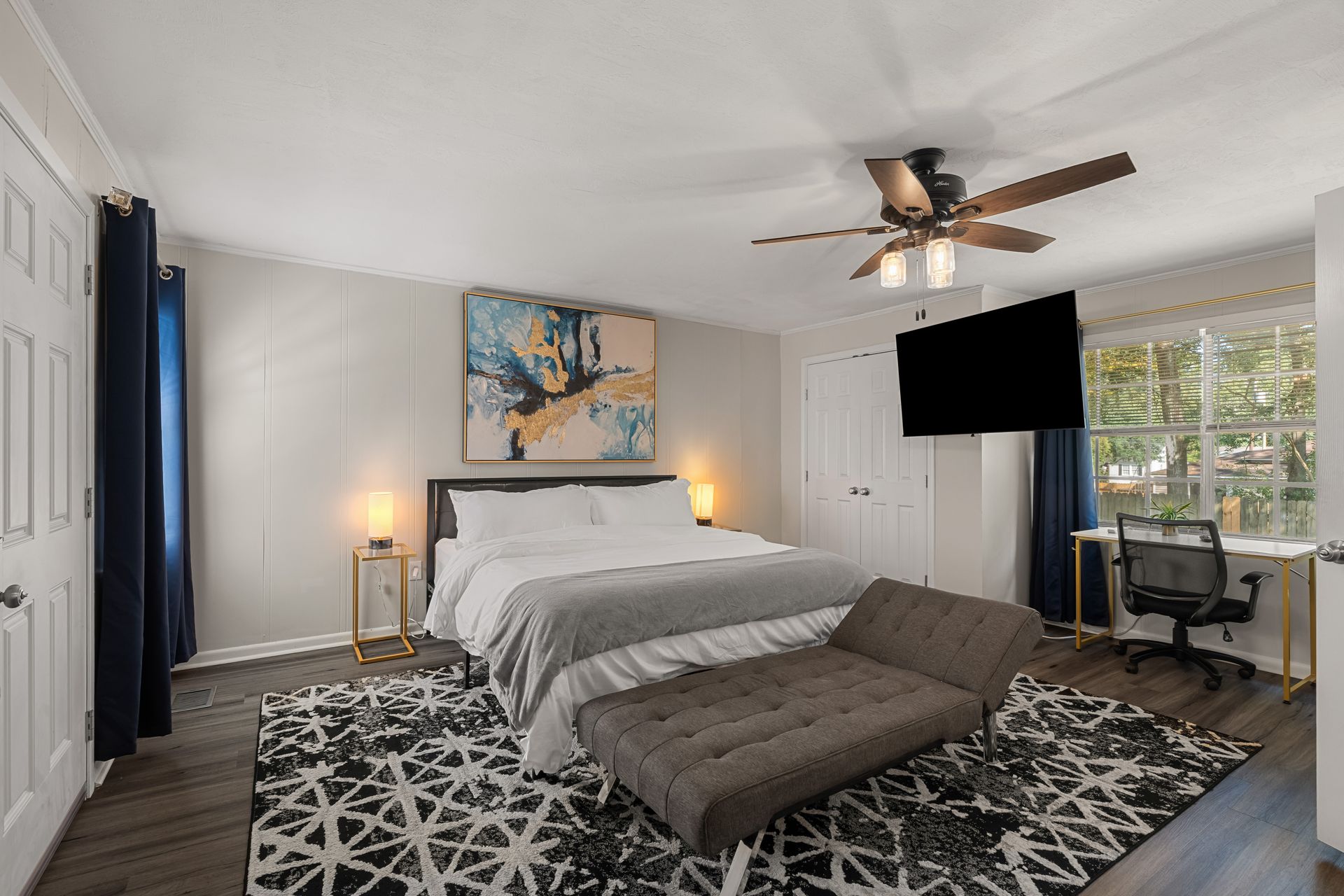 Bedroom with bed, art, rug, dark blue curtains, a desk, and a television.