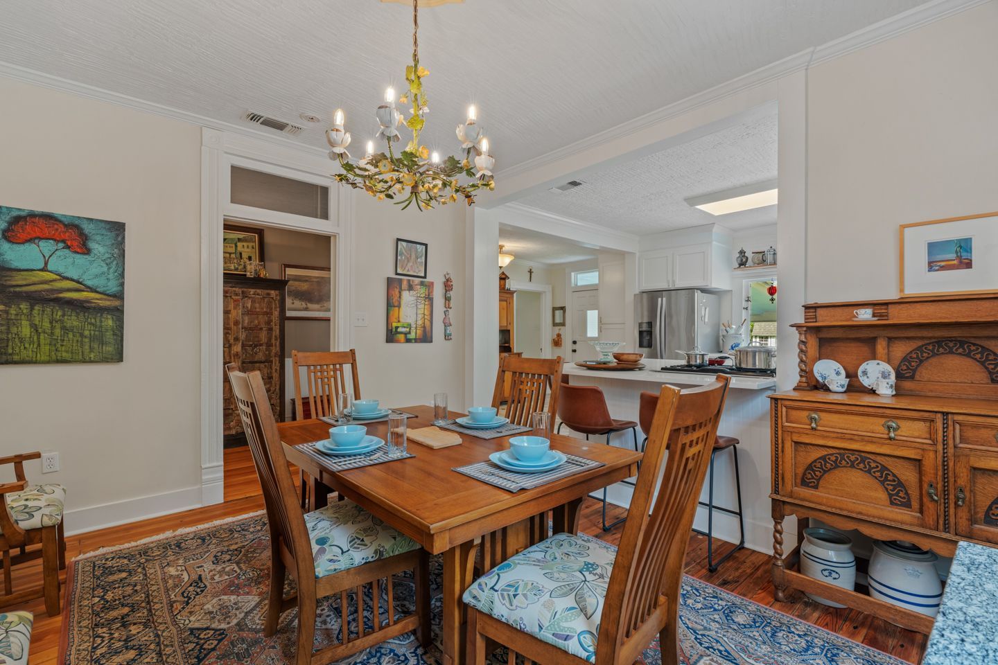 Dining room with wooden table, chairs, chandelier, and a view to the kitchen with a counter and stools.