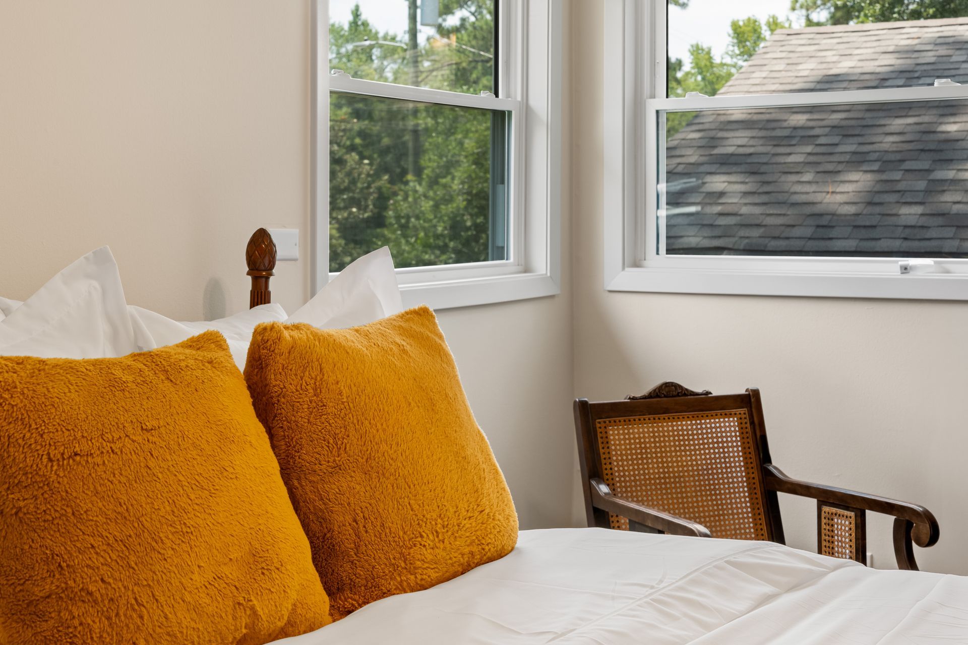 Bedroom corner with bed, orange pillows, and a chair near a window.
