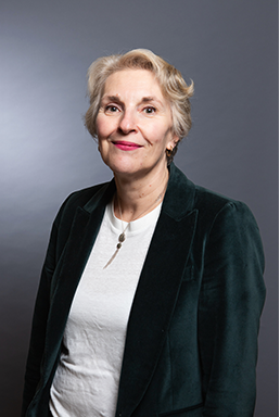 Une femme vêtue d'un blazer turquoise, d'une chemise blanche et d'un collier, sourit à l'objectif sur fond gris.