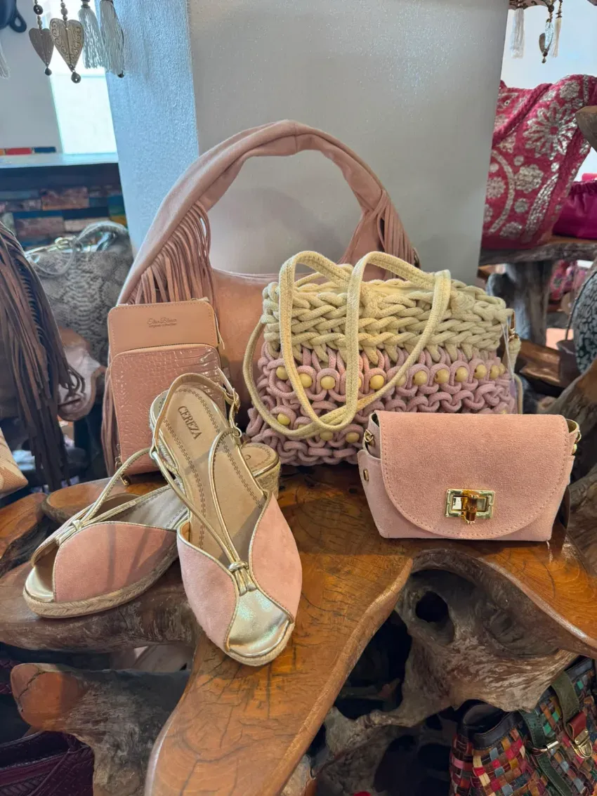 Varios bolsos y zapatos dispuestos sobre una mesa de madera junto a una cesta de mimbre y un monedero rosa.