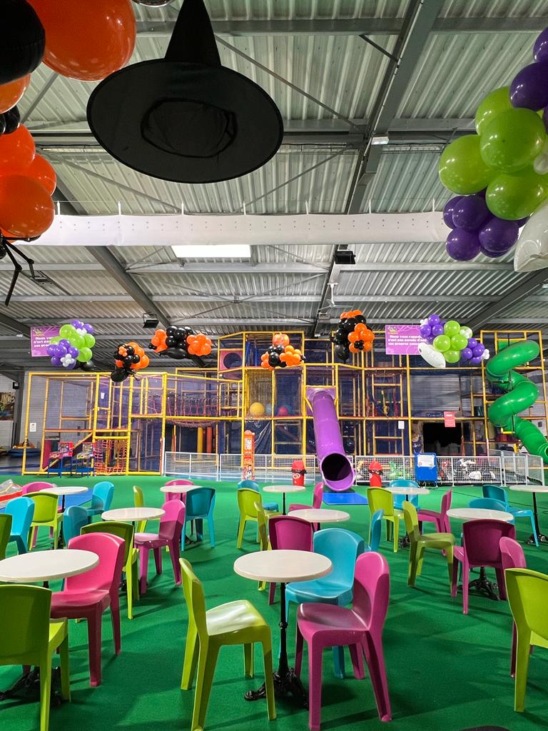 Aire de jeux intérieure décorée pour Halloween, avec tables, chaises et structure de jeux. Des ballons et un chapeau de sorcière sont suspendus au plafond.