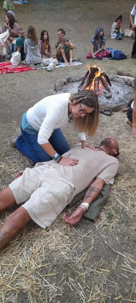 Mujer realizando RCP a una persona tendida en el suelo, con una fogata y un grupo de personas al fondo.