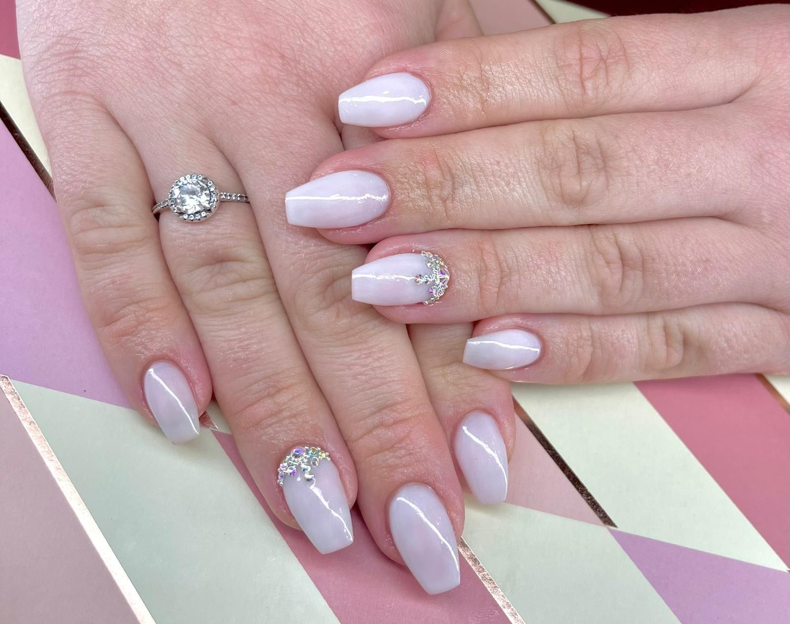 Mãos com unhas rosa claro, em formato oval, algumas decoradas com cristais, e um anel no dedo.