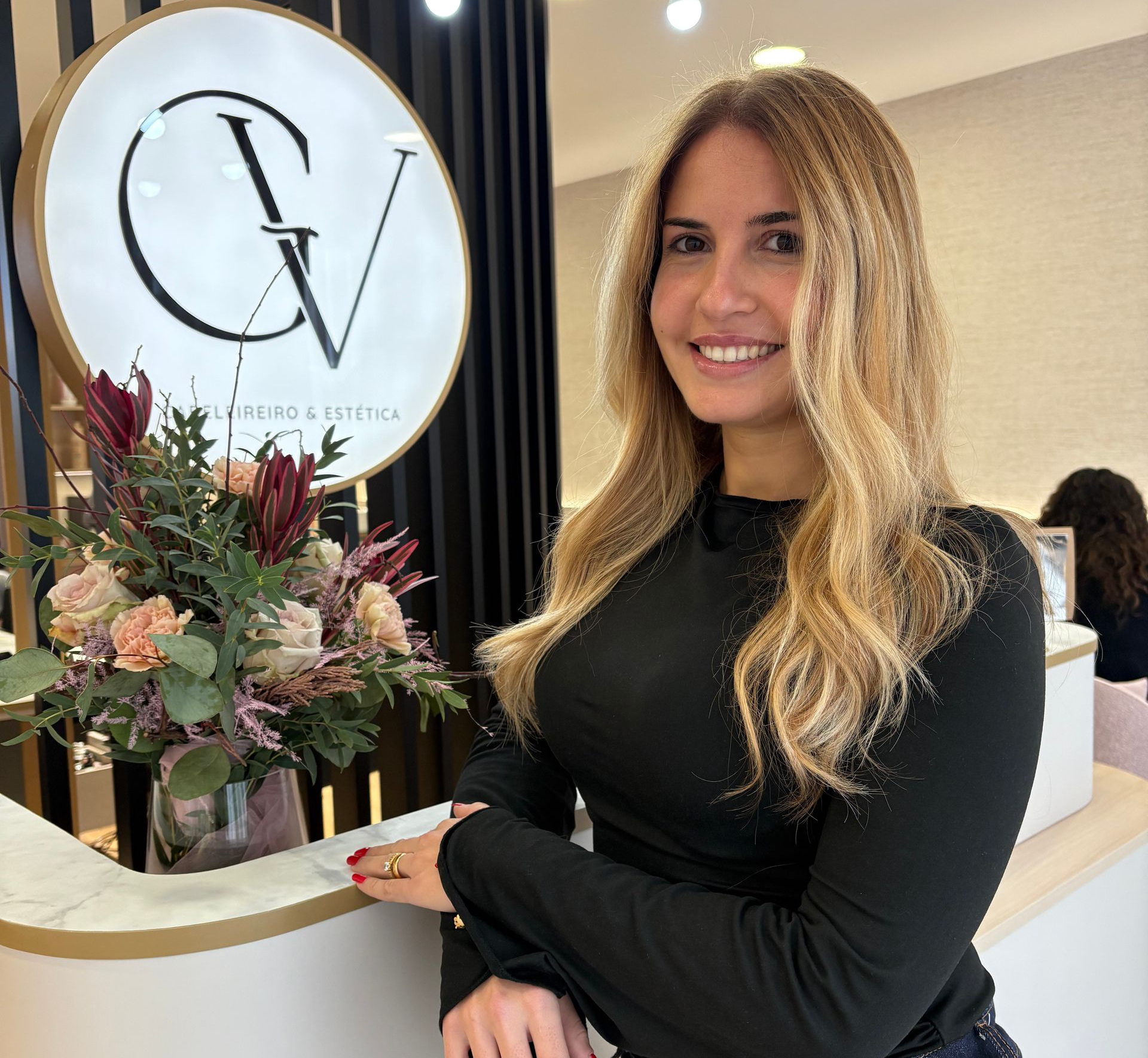 Mulher sorrindo, posando ao lado do logotipo de um salão de beleza; flores ao fundo.
