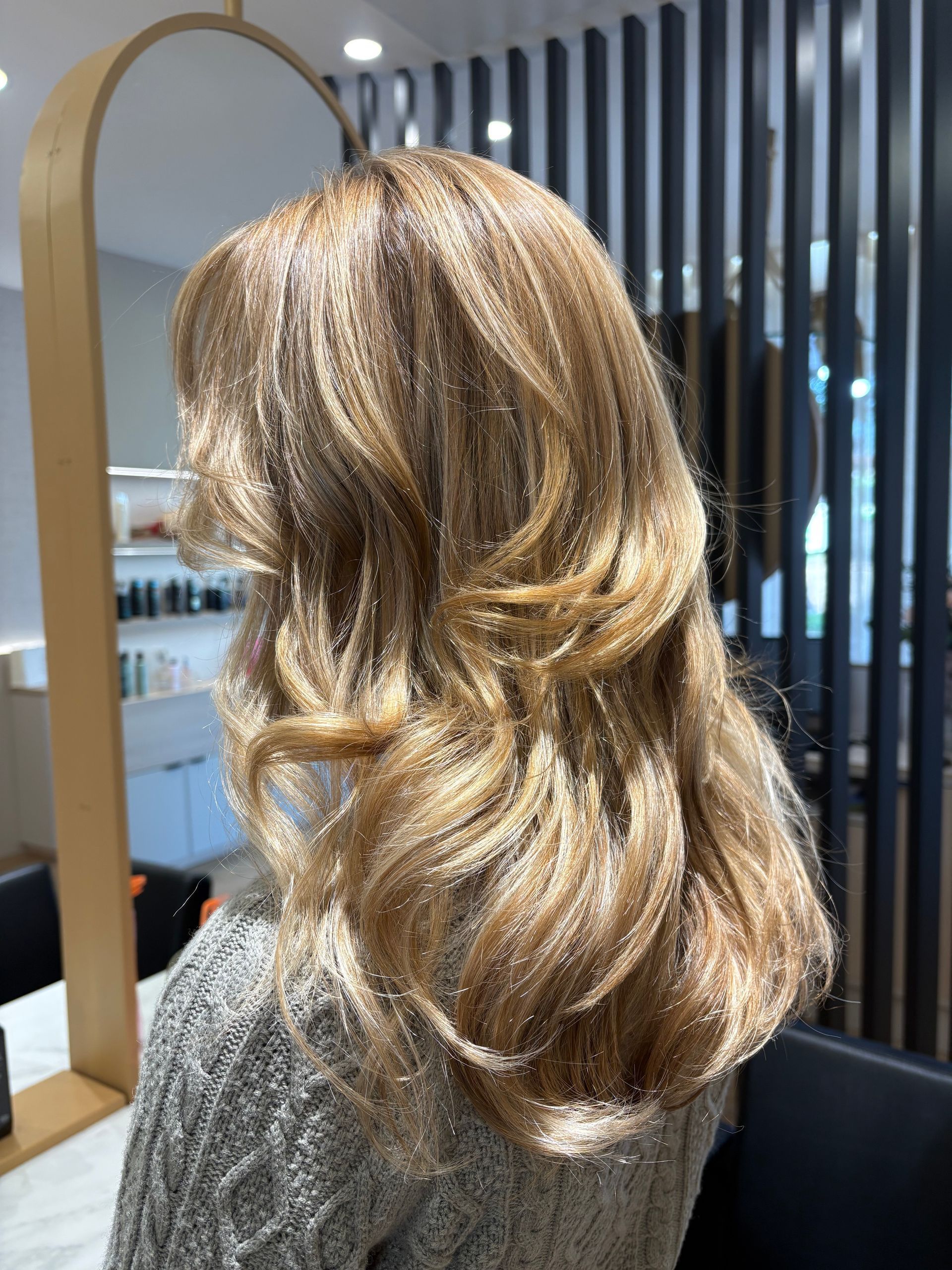 Mulher com cabelo castanho-dourado ondulado e em camadas, estilizado em salão de beleza com espelho e painel ripado ao fundo.