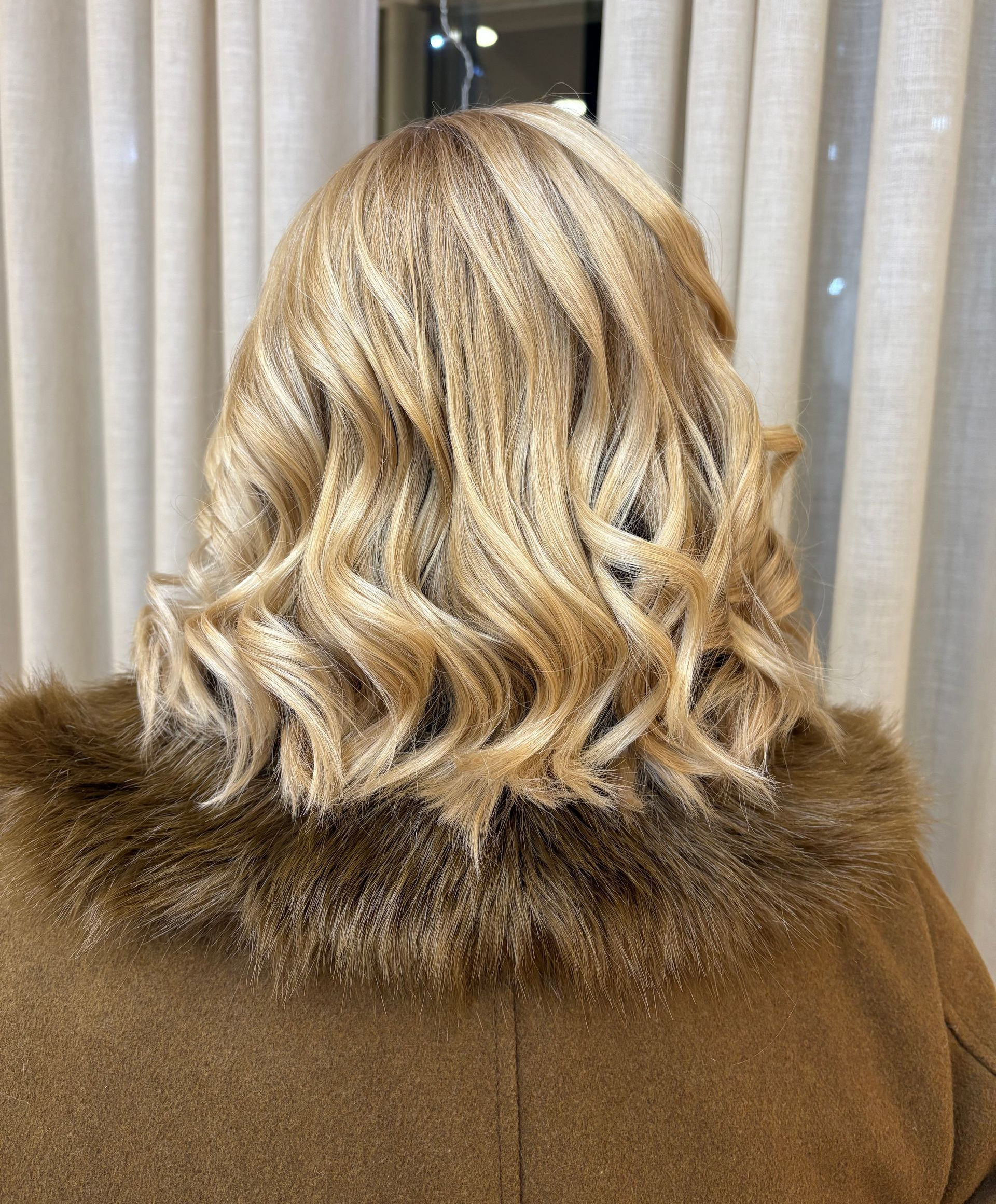 Cabelo loiro ondulado estilizado com uma gola de pele marrom sobre um casaco marrom.
