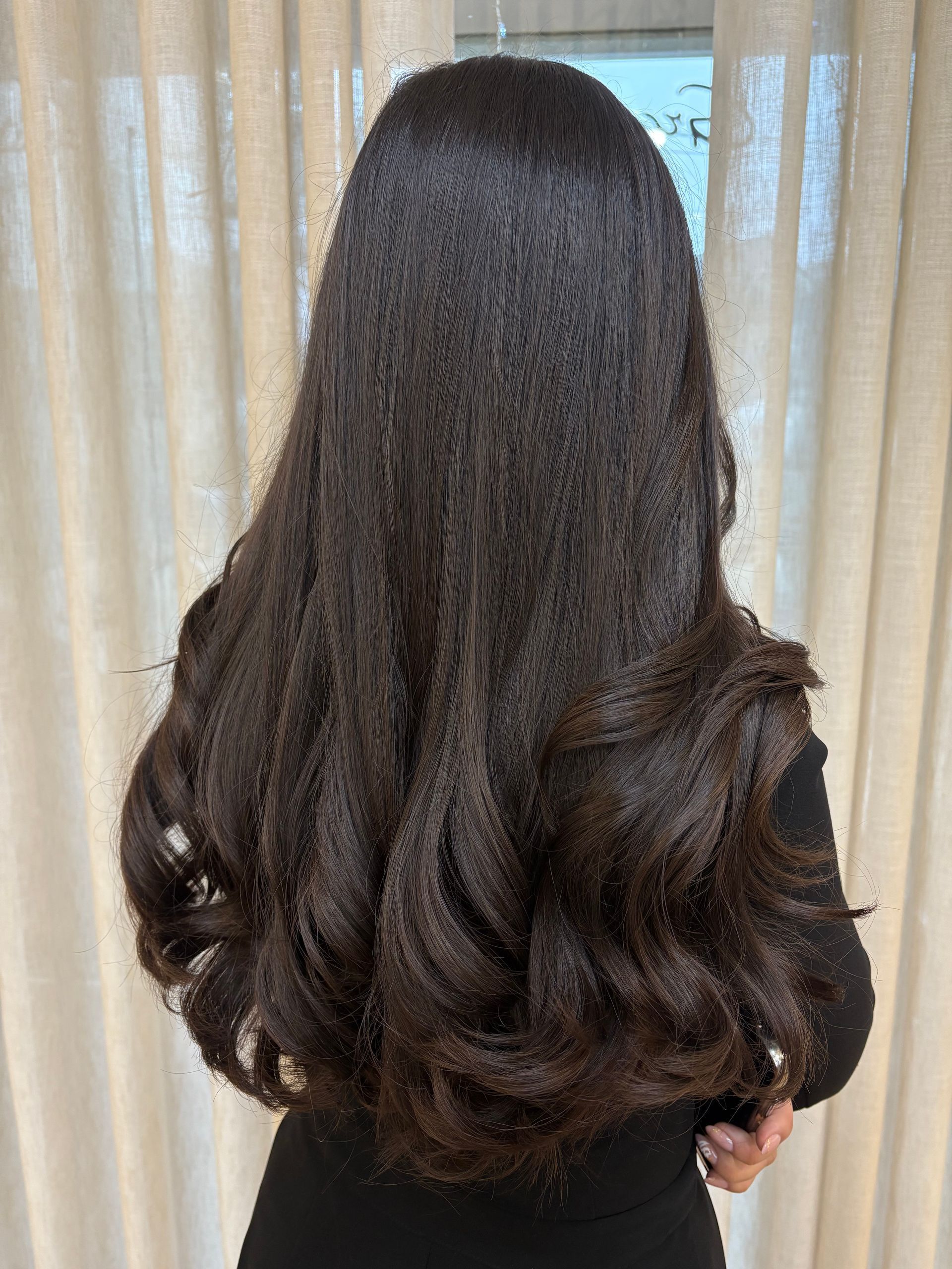 Mulher com cabelos longos, ondulados e escuros. Vista de costas, em ambiente interno, vestindo uma blusa preta.