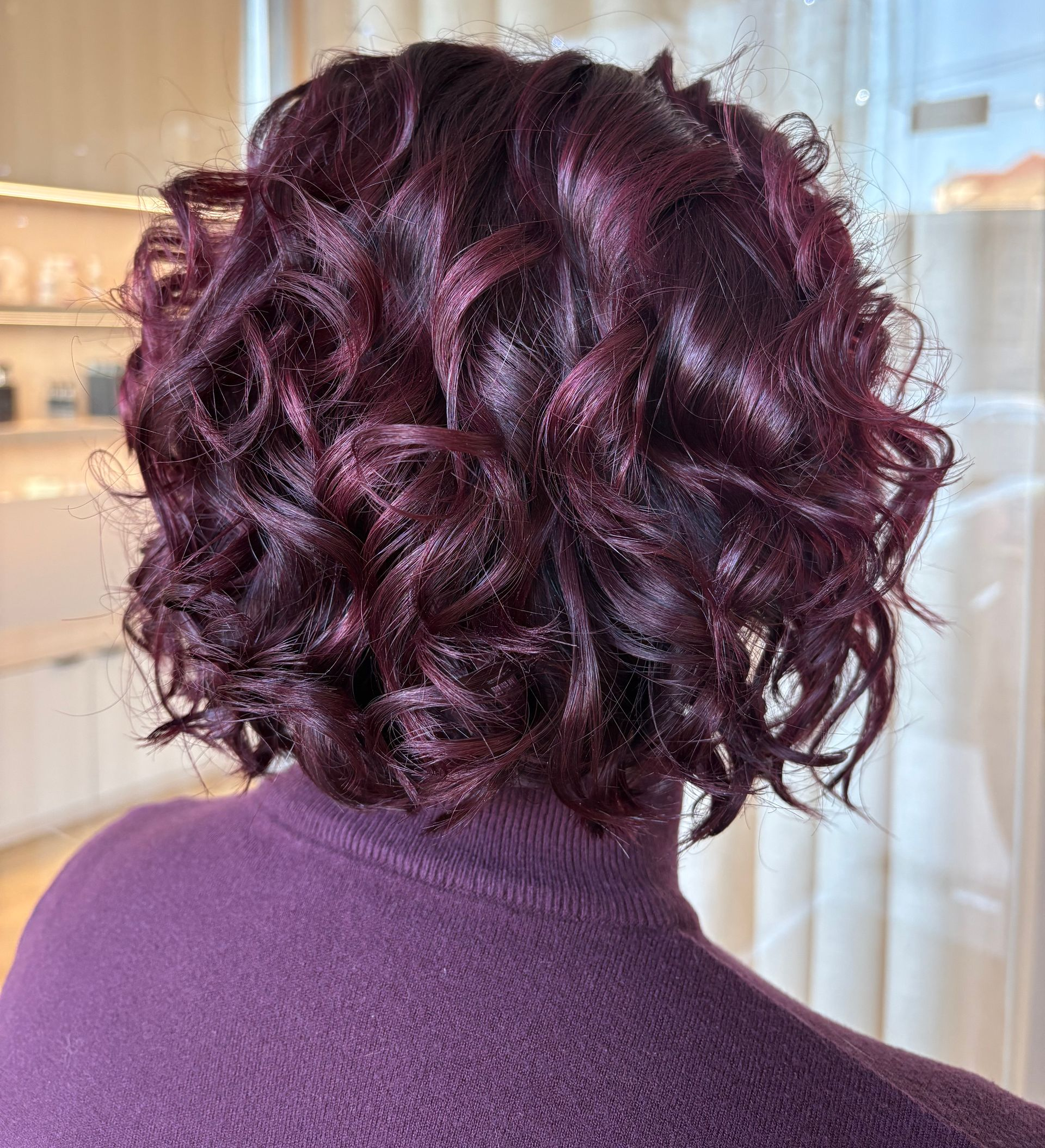 Pessoa com cabelo encaracolado cor de vinho e uma blusa de gola alta roxa combinando, em ambiente interno.