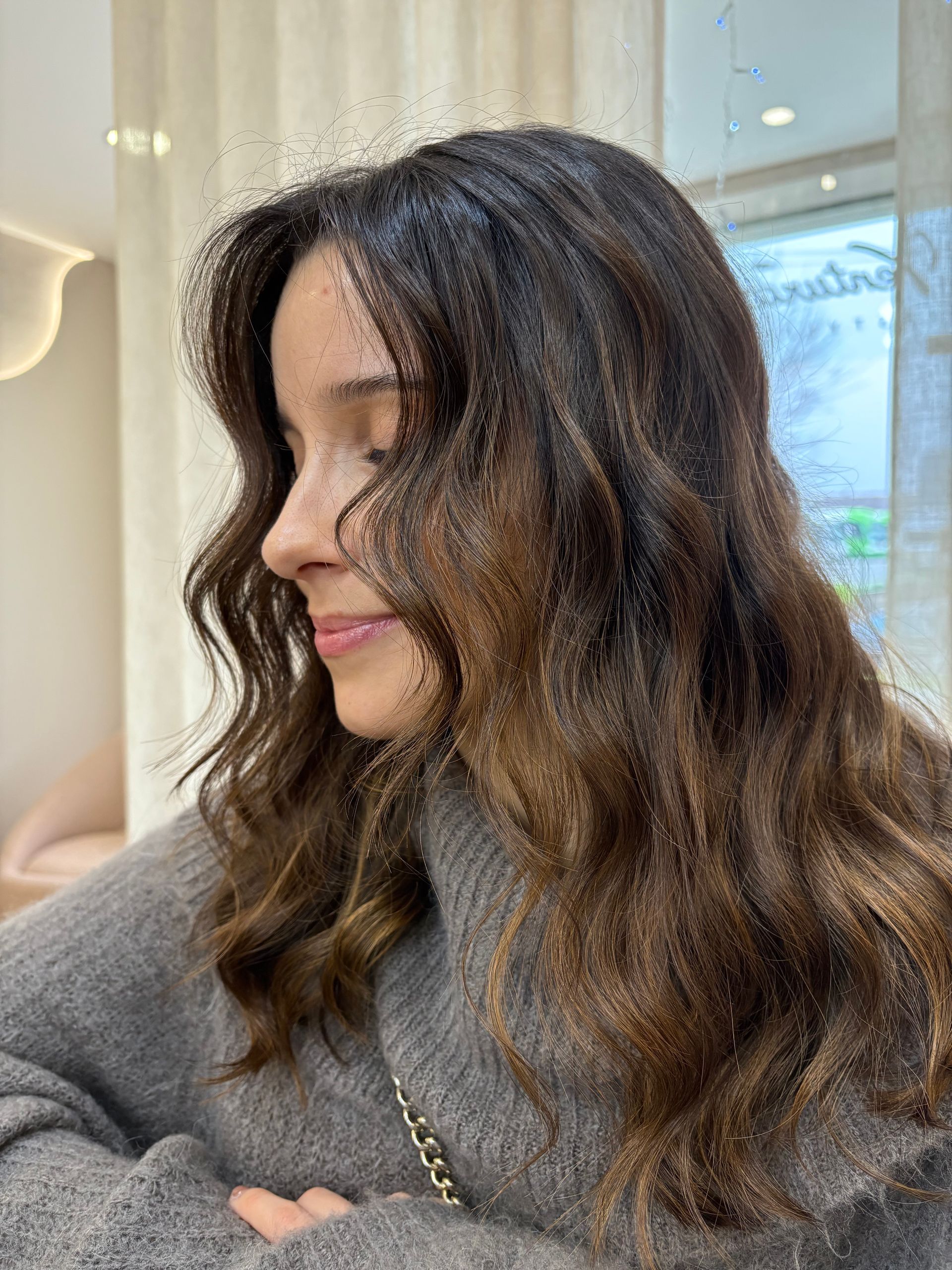 Mulher de cabelos castanhos ondulados, vestindo um suéter cinza, olhos fechados, sorrindo, em ambiente interno.