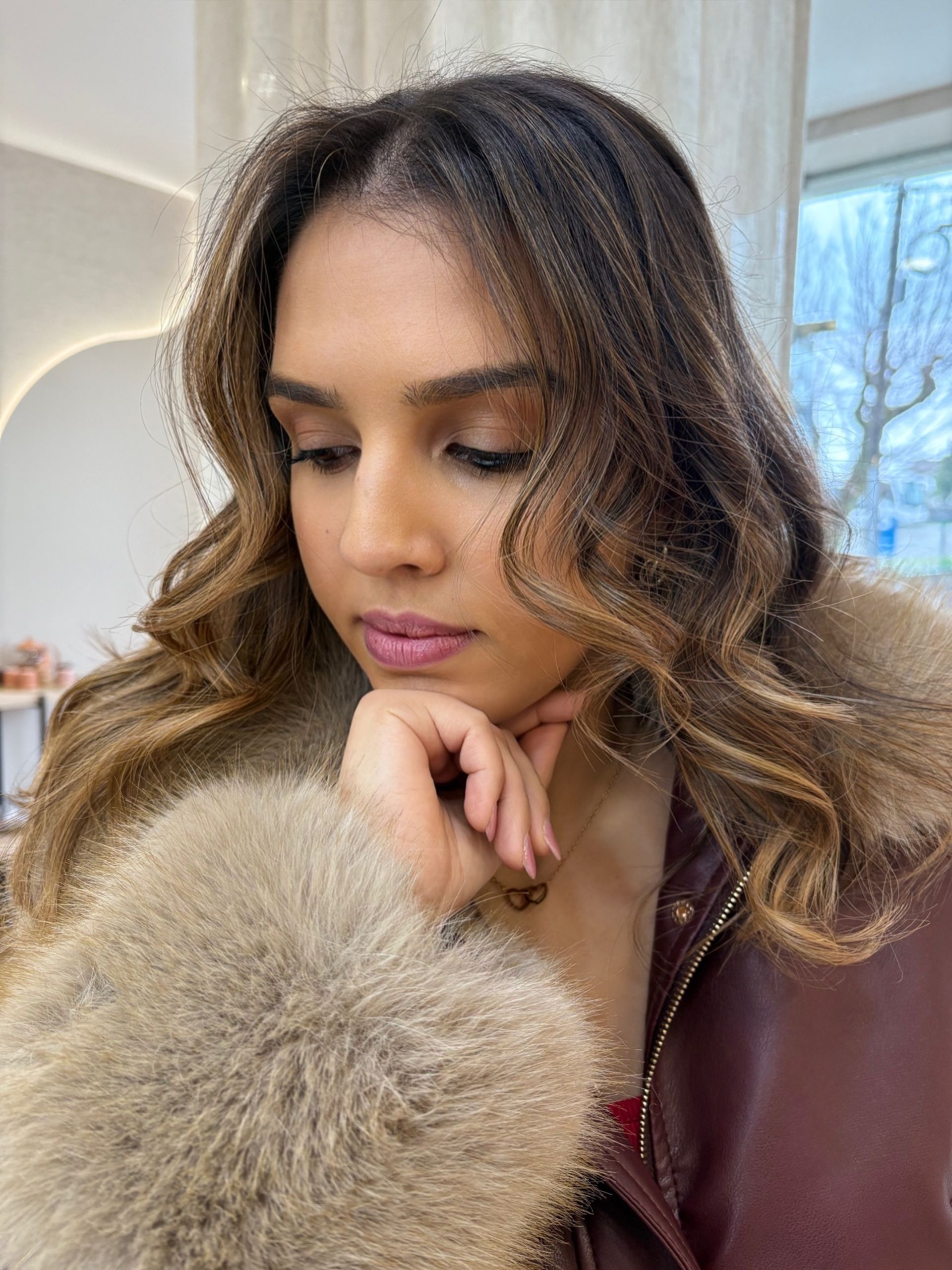 Mulher de cabelos castanhos ondulados, com o queixo apoiado na mão, vestindo jaqueta com detalhes em pele