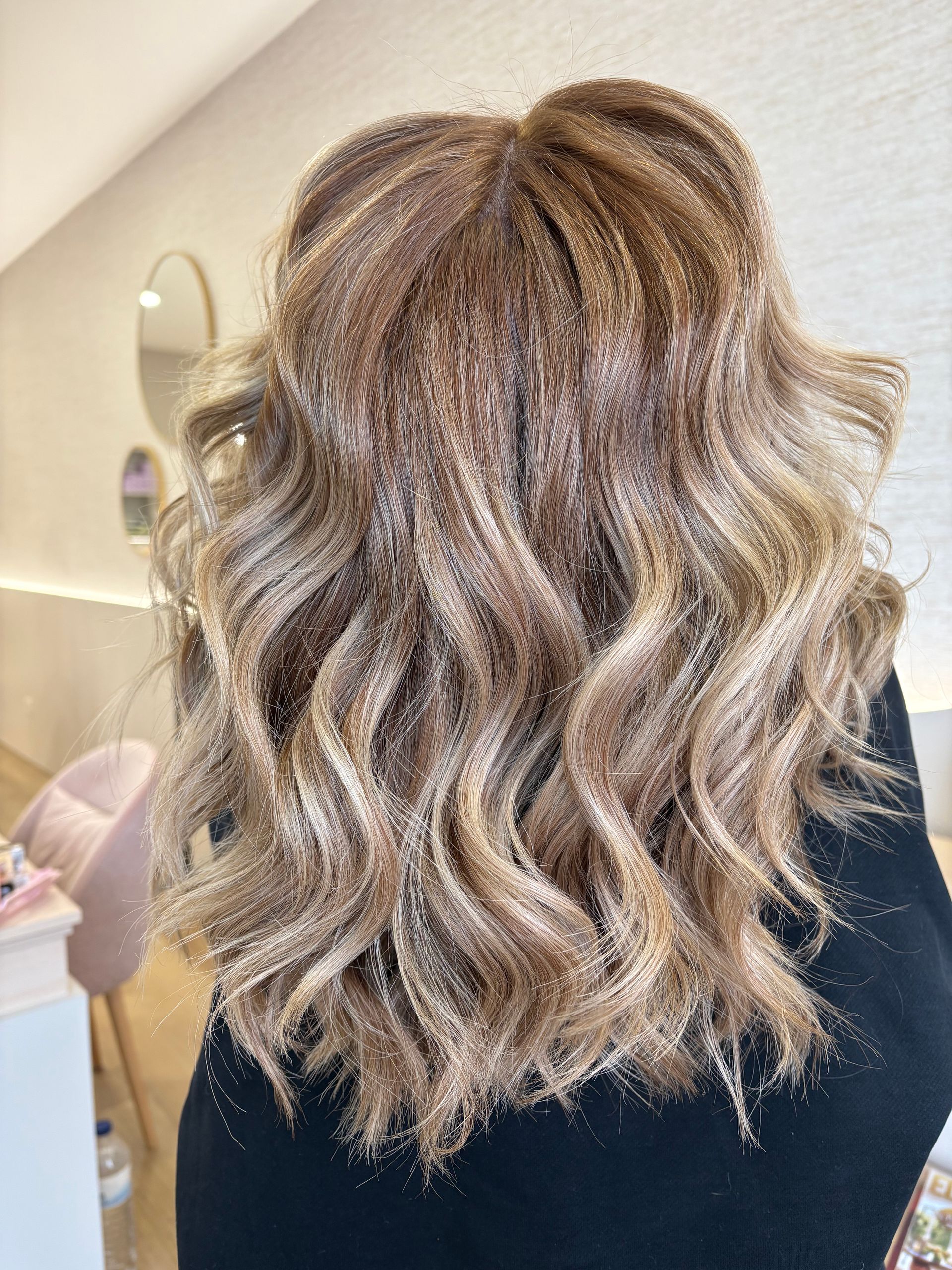 Cabelo loiro ondulado com mechas brilhantes, efeito ombré, repousando nas costas de uma pessoa; blusa listrada azul.