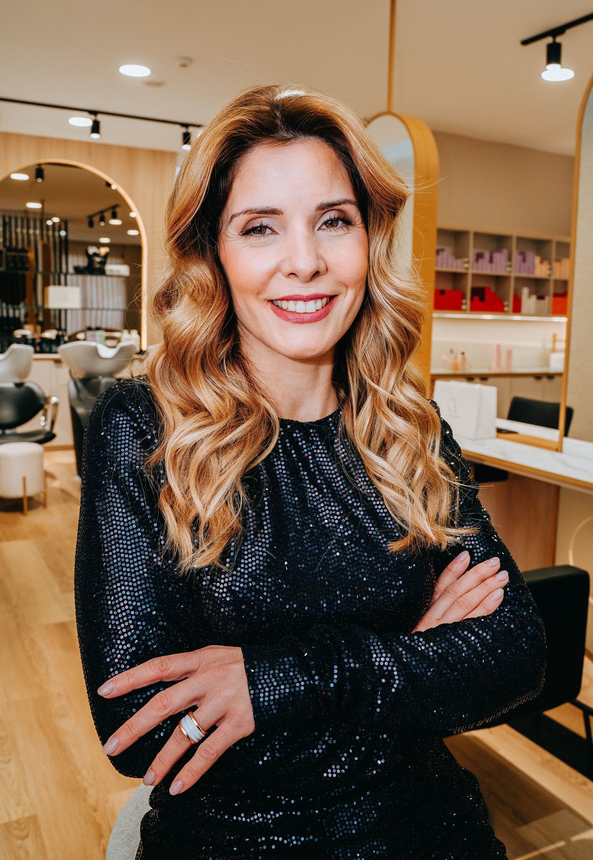 Mulher com vestido de lantejoulas sorri, de braços cruzados, em um salão de cabeleireiro.