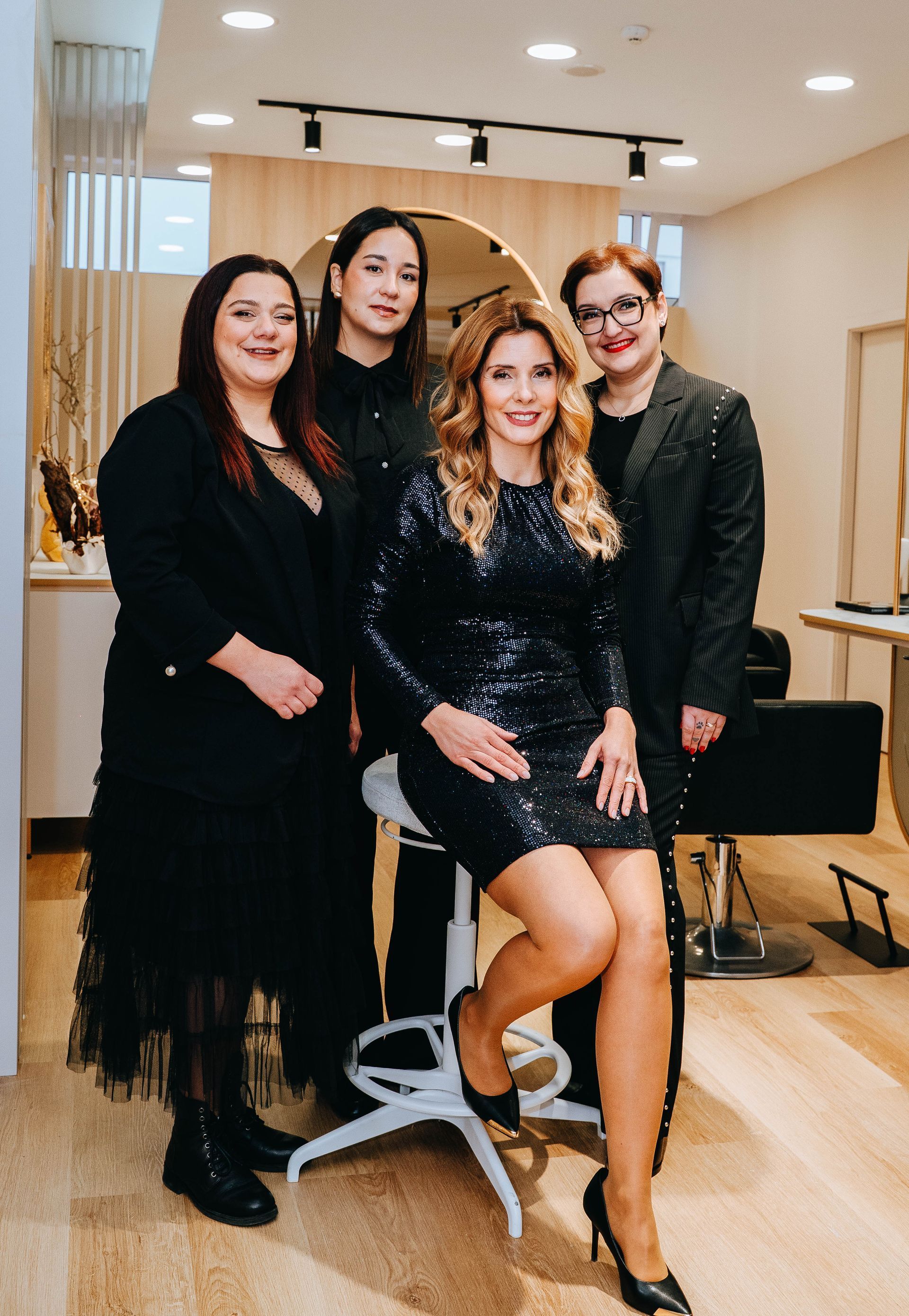 Quatro mulheres posam em um salão de cabeleireiro. Uma está sentada, usando um vestido de lantejoulas.