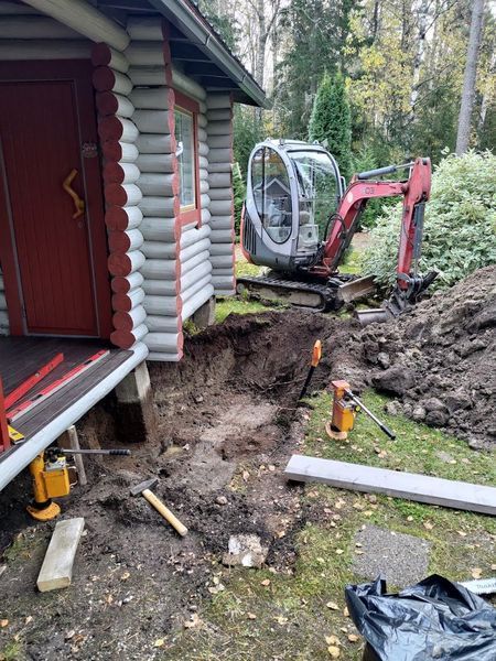 Minikaivuri kaivaa ojaa hirsimökin vieressä, maa-kasojen ollessa perustusta pitkin.