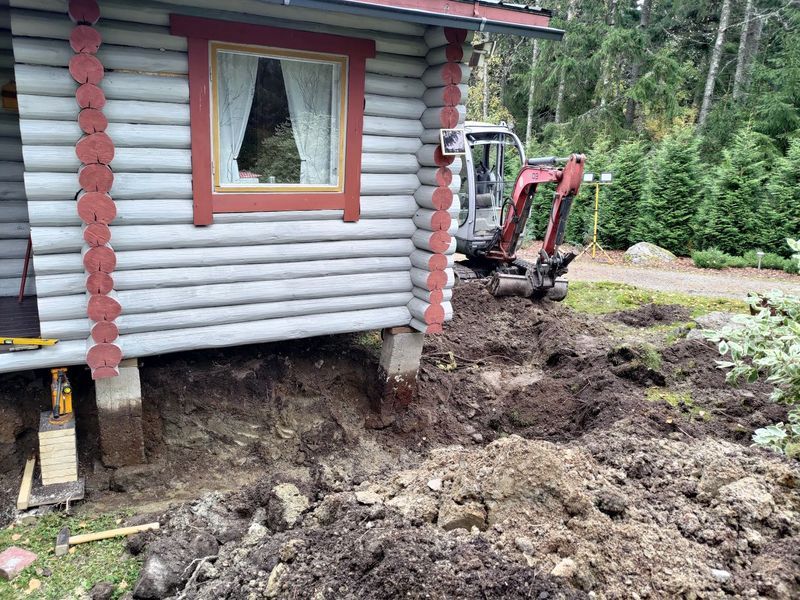Mökin ulkopinta, kaivinkone kaivaa mutaa perustusten ympärillä metsäisellä pihalla