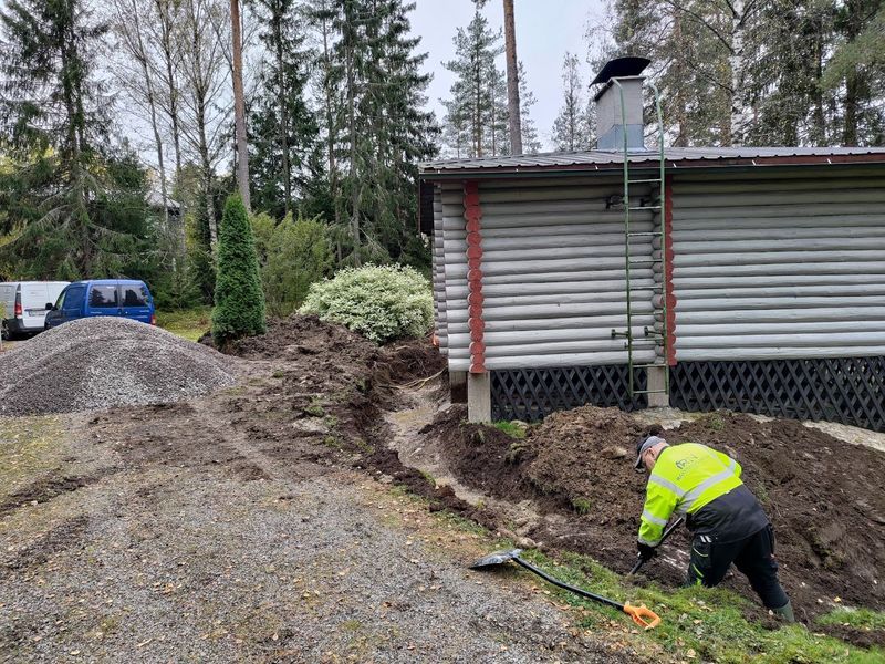 Neonväriseen takkiin pukeutunut työntekijä kaivaa ojaa vajan vieressä sorapihalla.