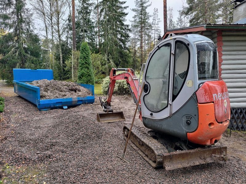 Pieni oranssi minikaivuri pysäköitynä sinisen roskalavan viereen metsäisellä työmaalla