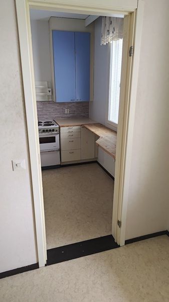 Näkymä oviaukosta pieneen keittiöön, jossa on siniset kaapit, liesi, pesuallas ja ikkuna.